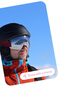Man wearing a black helmet and reflective ski goggles with snowy mountains reflected, dressed in an orange and blue jacket against a clear blue sky.